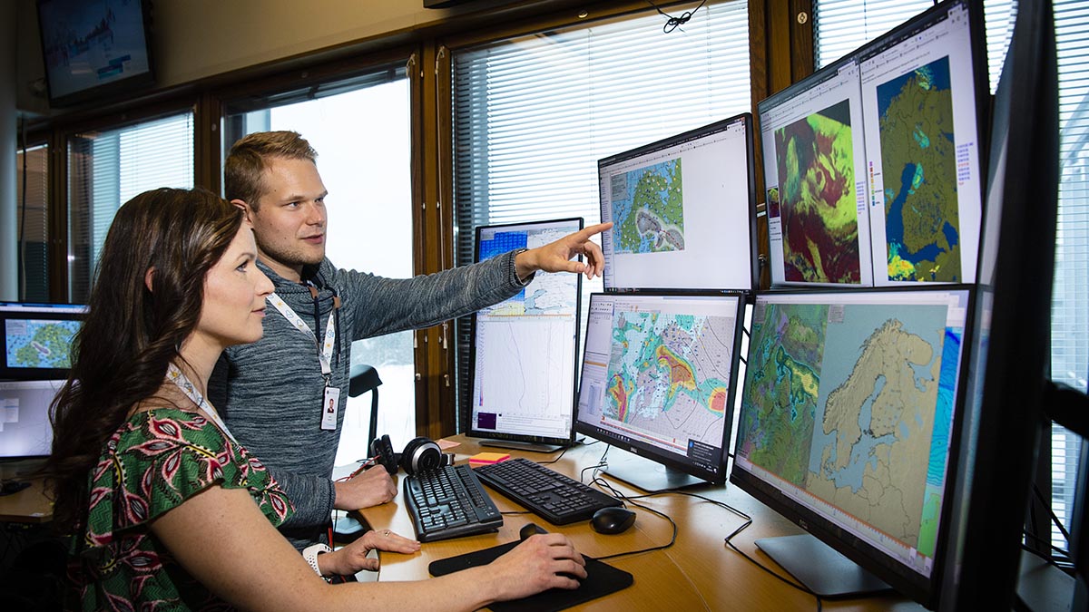 Two meteorologists forecasting the weather. Several monitors in front.
